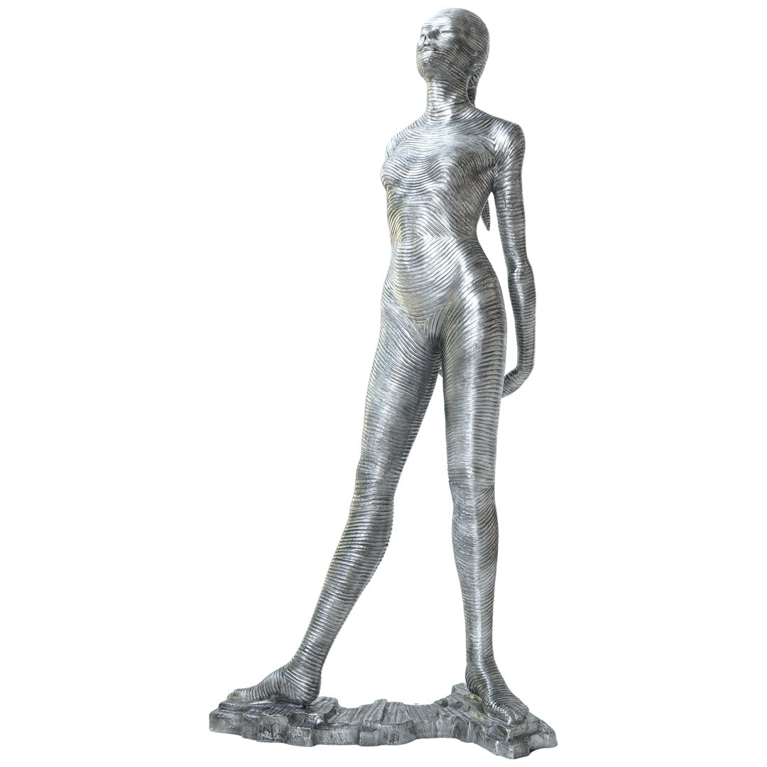 A metallic silver sculpture of a standing female figure with a textured, ribbed surface, mounted on a rocky base against a plain white background.