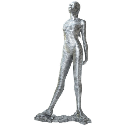 A metallic silver sculpture of a standing female figure with a textured, ribbed surface, mounted on a rocky base against a plain white background.
